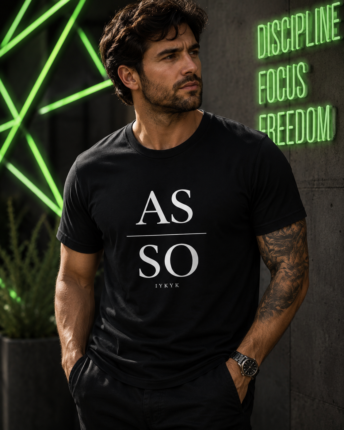 Man wearing black As Above So Below IYKYK t-shirt in a dark setting with green light and discipline focus freedom wall text.