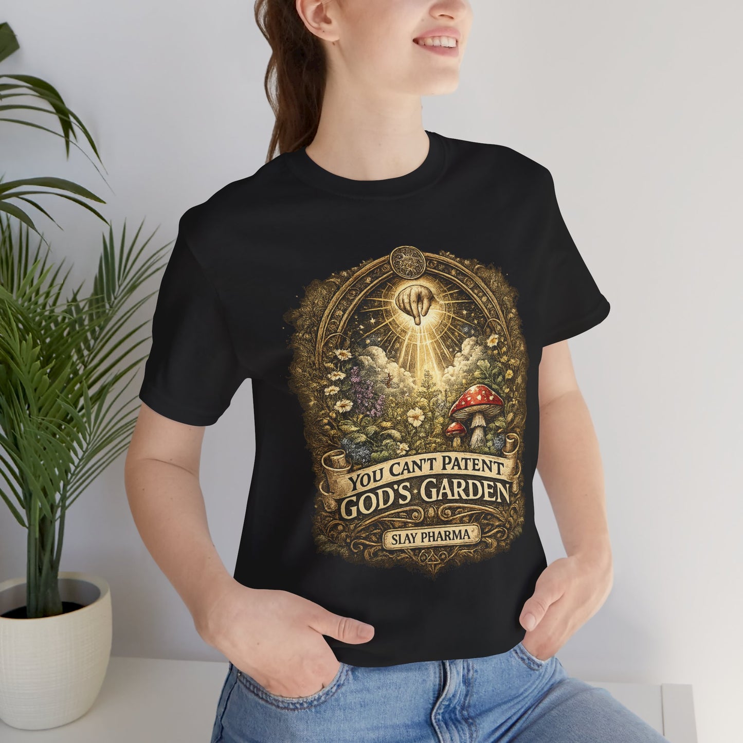 Woman wearing You Can’t Patent God’s Garden black t-shirt indoors near a plant, showing the sacred botanical design.